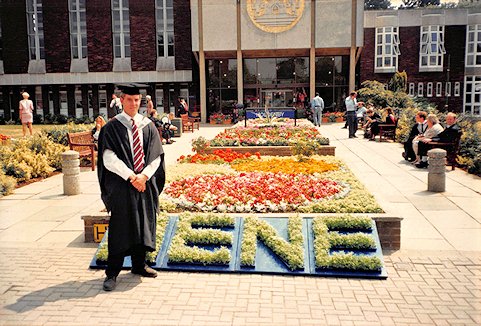 A graduate standings next to flower beds, including 'Nene' in topiary